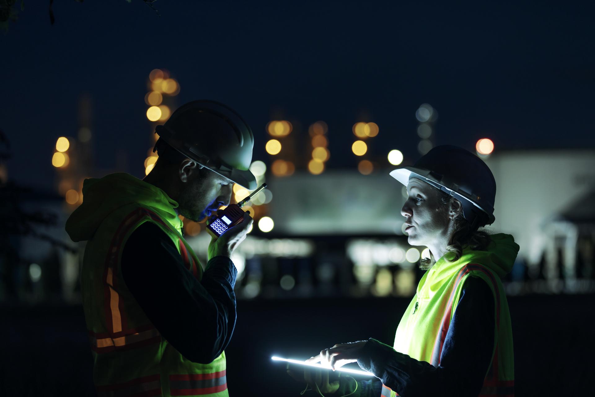 Two engineers in hard hats reviewing a tablet on site at night